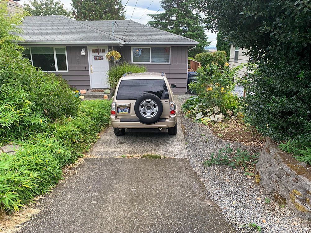 Tan SUV parked in driveway of a one-story house with greenery on either side.