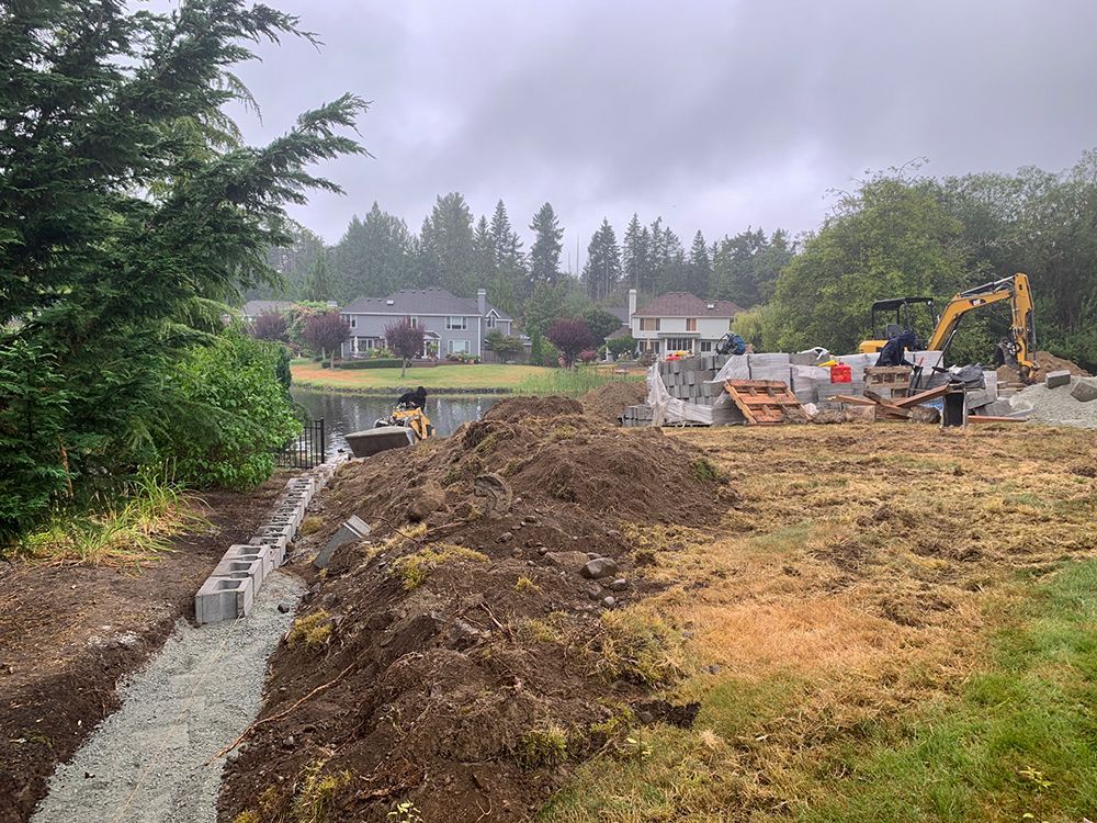 Construction site near a body of water with an excavator, blocks, and dirt. Houses in the background on a cloudy day.