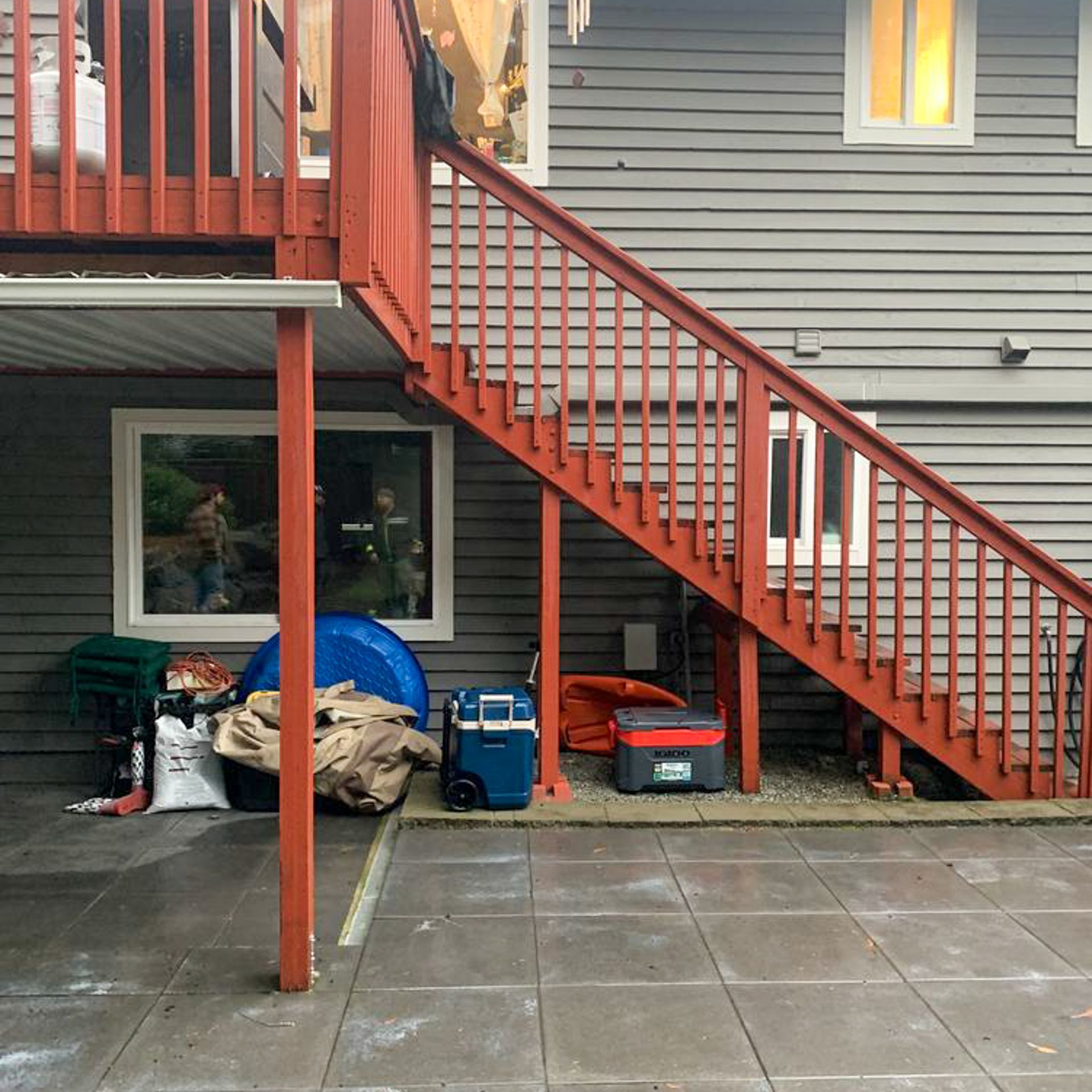 Red stairs lead to a deck. Underneath, gray house siding, storage, and windows.