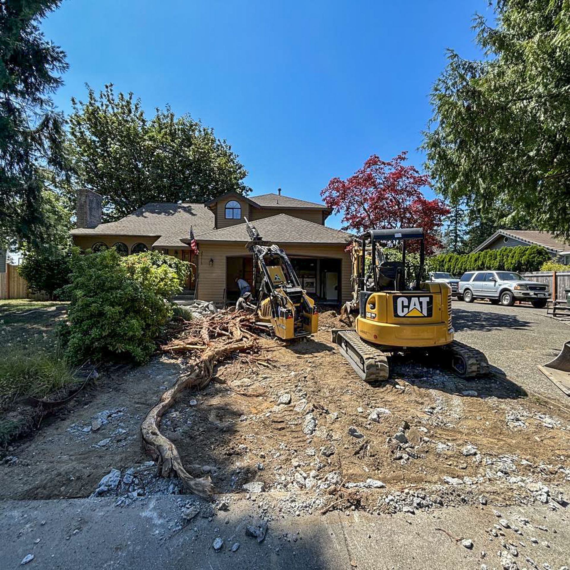Construction site: Two yellow excavators in front of a house, removing tree roots under a blue sky.
