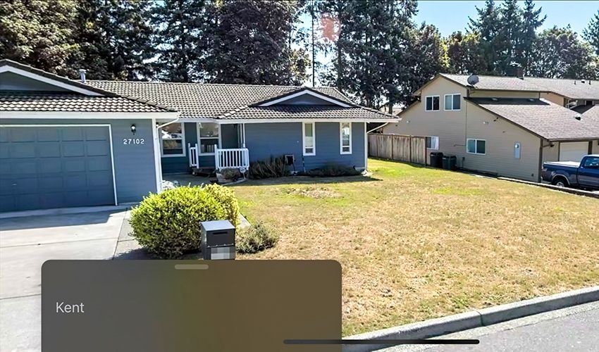 A blue house with a green lawn and a gray garage; in the background, more houses and trees.