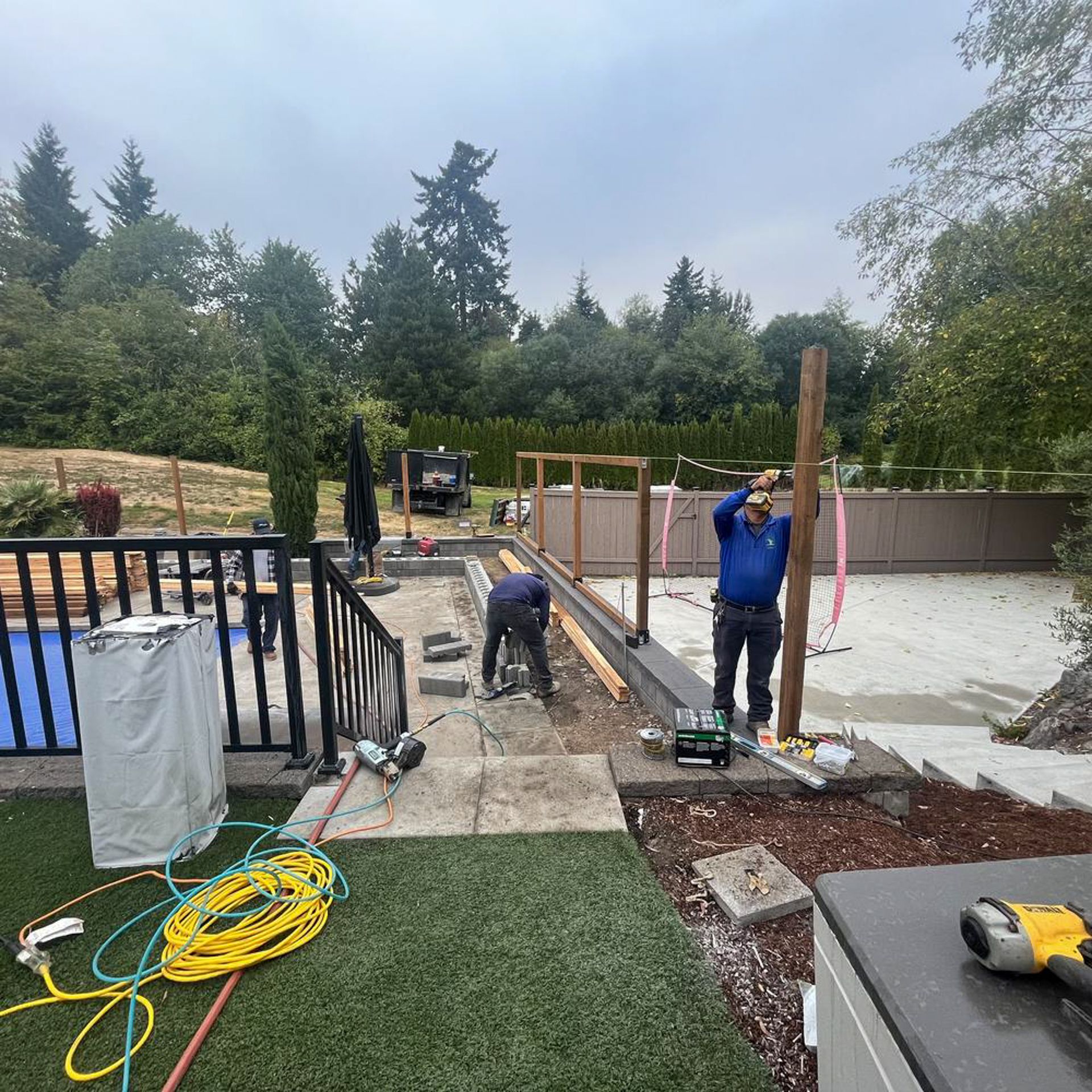Two men build a wooden structure outside. One man drills a post, the other works on the ground near steps. Overcast day.