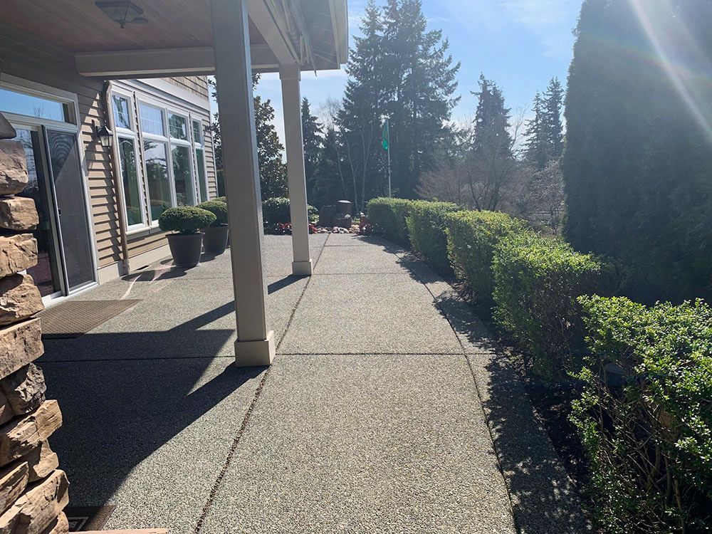Concrete walkway with bushes on right, porch with columns on the left, and trees in the background.
