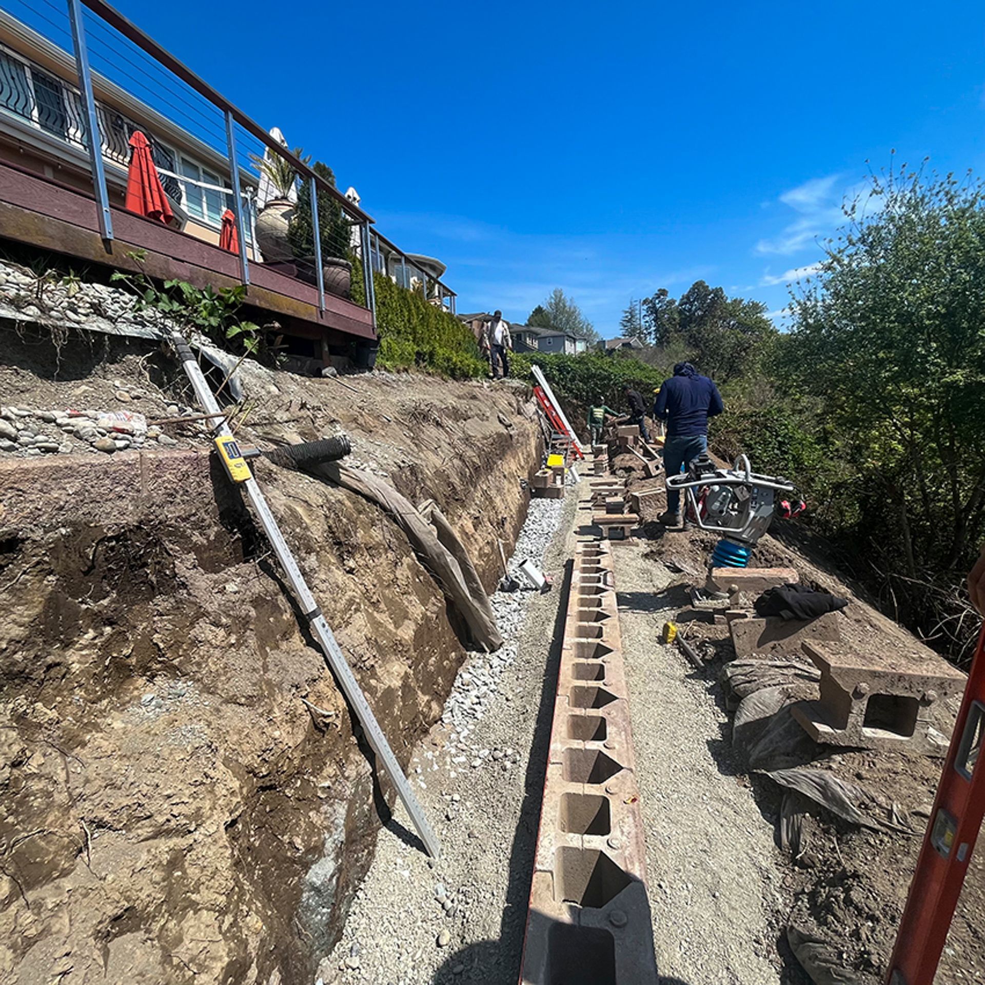 Construction of a retaining wall; workers in a trench, using cinder blocks and tools, with a house on a hillside.