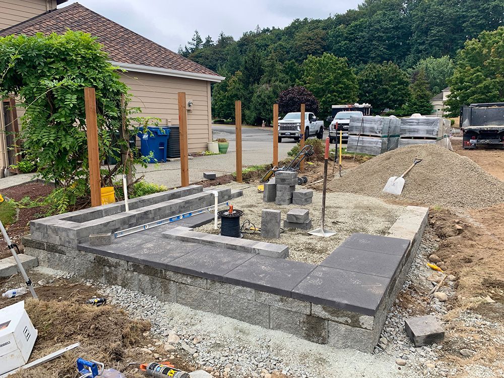 A raised garden bed under construction with gray blocks, tools, and a building in the background.