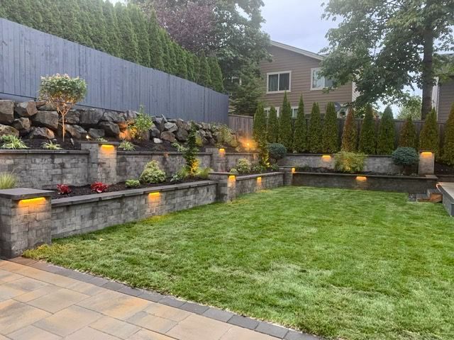 A terraced backyard with retaining walls, lighting, green grass, and plantings, backed by a fence and trees.