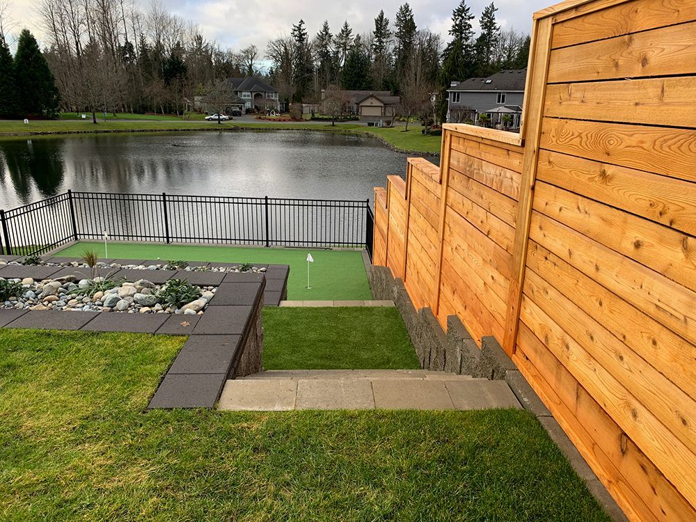 A backyard with a putting green, lake, and wooden fence. Green grass and stone steps.