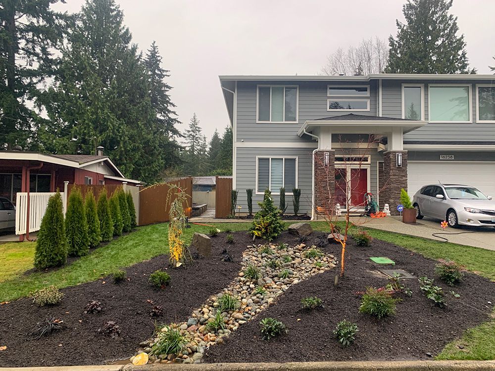 Two-story gray house with new landscaping, including a rock garden, lawn, and evergreen trees.