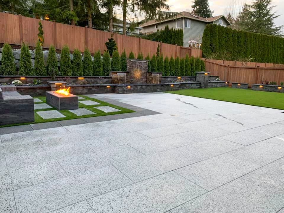 Backyard patio with fire pit, water feature, and grass lawn; wood fence and greenery in the background.