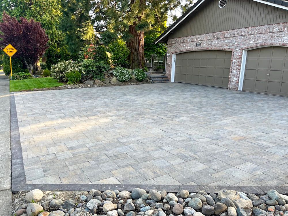 Paver driveway in front of a house with a two-car garage. Landscaping and a road sign are visible.