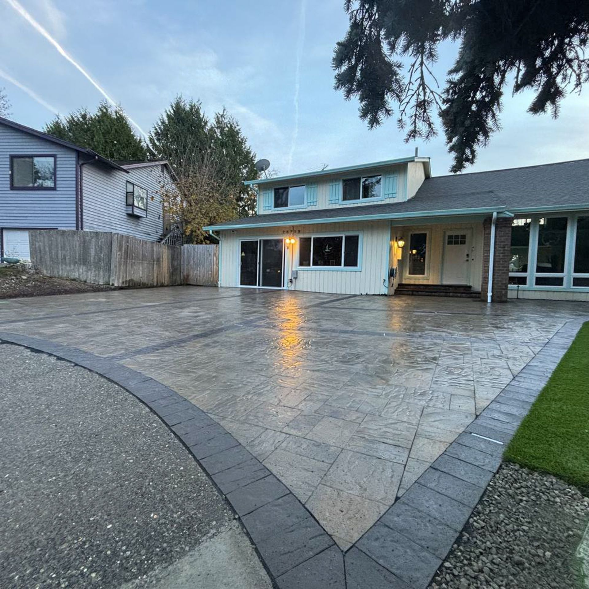 Front view of a house with a concrete driveway, bordered by dark gray paving stones, under a cloudy sky.