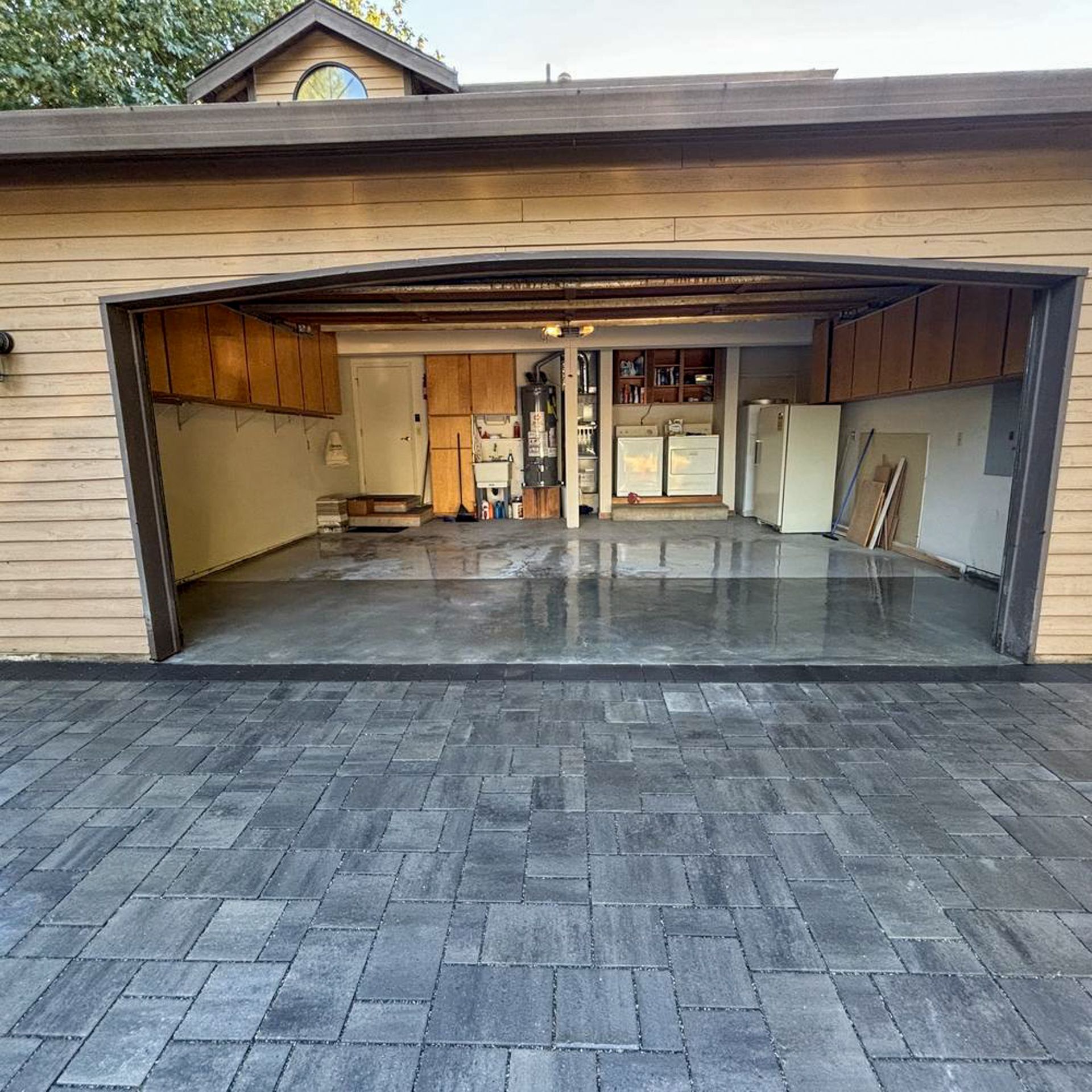 Garage entrance with newly paved gray brick driveway. Inside, a partially empty garage with cabinets and appliances.