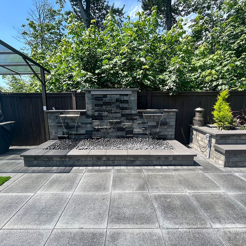 Water feature with stone facade in a paved backyard. Water cascades over dark stones into a bed of black rocks.