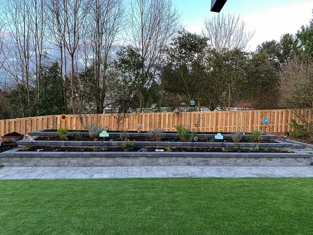 Raised garden beds with plants, bordered by stone, in a backyard with a wooden fence.