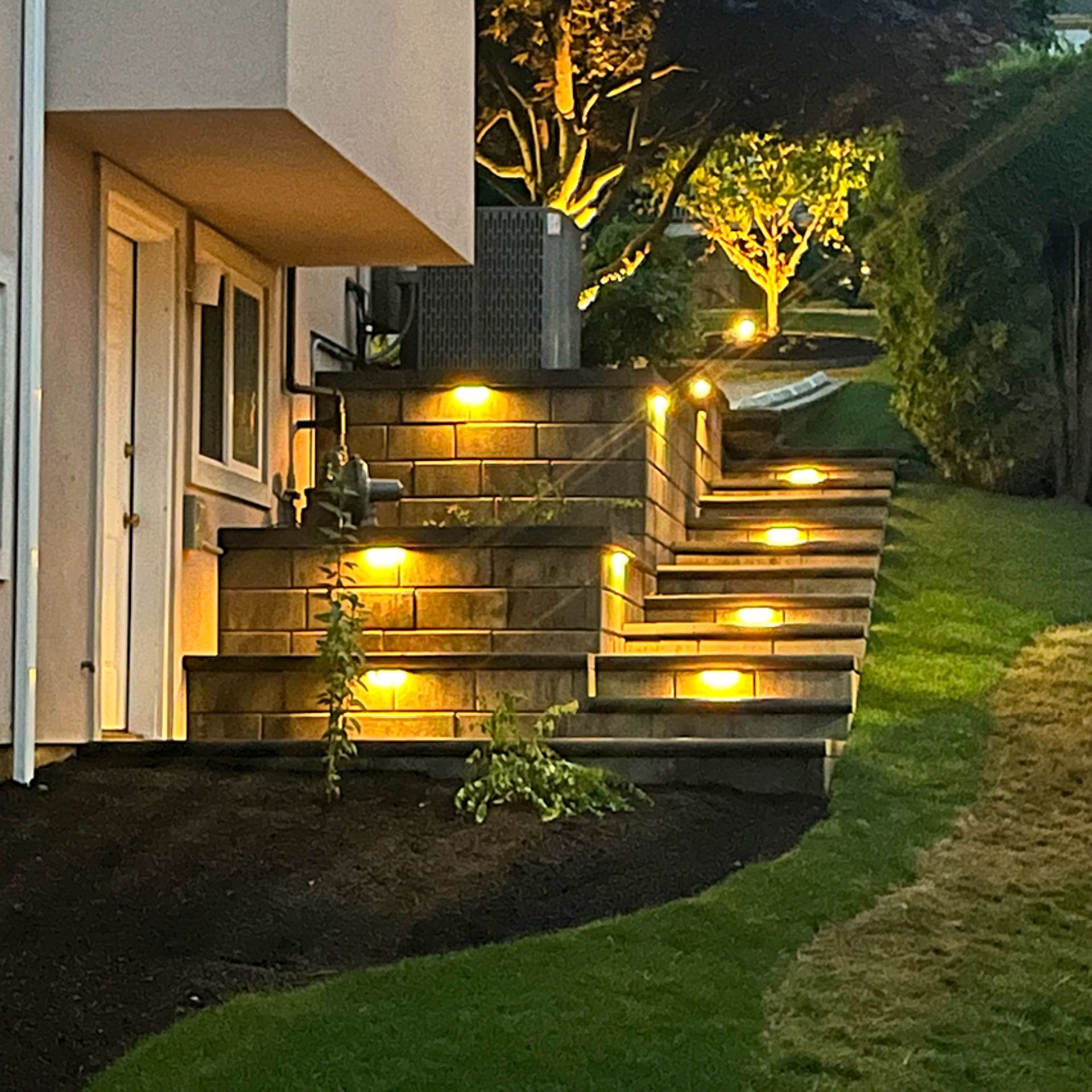 Lit stone steps lead up a hill at night, with landscape lighting and lawn.