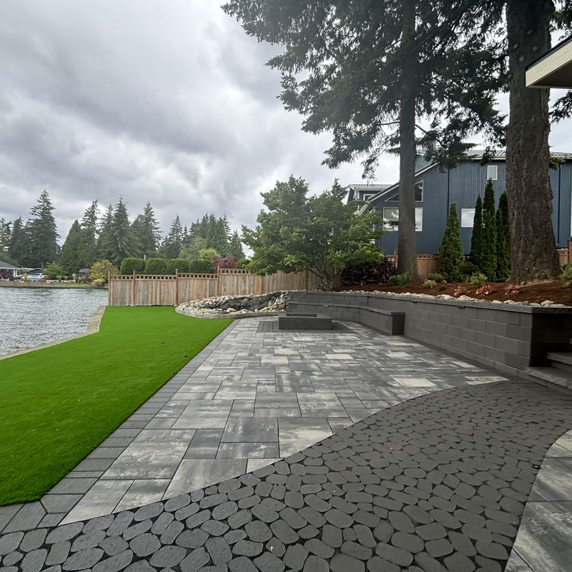 Patio by a lake with gray pavers, green turf, and a wooden fence on a cloudy day.