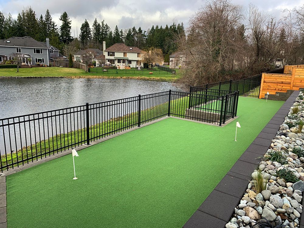 Artificial turf putting green with two holes, by a pond, fenced, with tiered wooden landscaping.