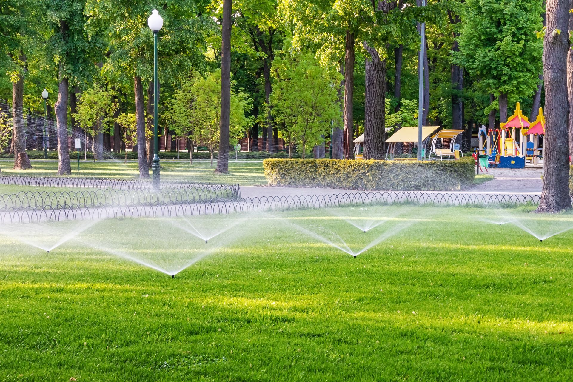 Sprinklers watering a green lawn in a park, with trees, a playground, and kiosks in the background.