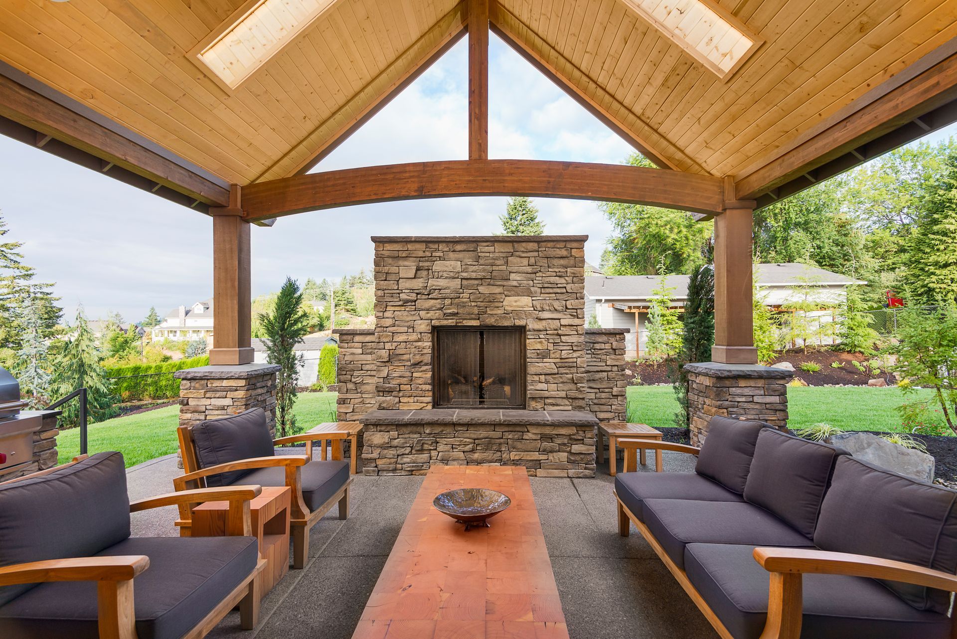 Outdoor patio with stone fireplace, wood furniture, and green landscaping.