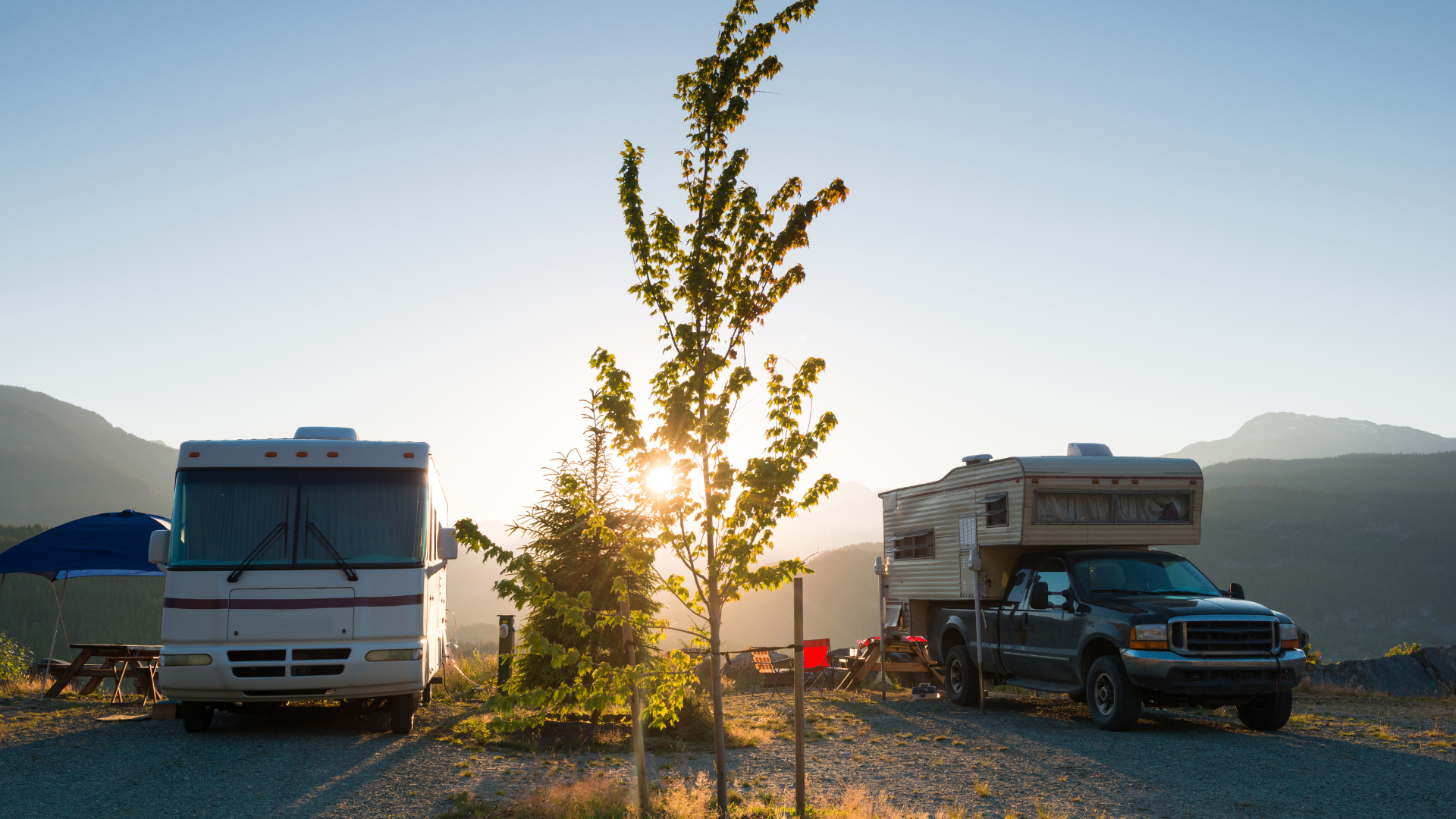 Two RVs parked at a campsite with a mountain backdrop. The sun shines through a small tree in the center.