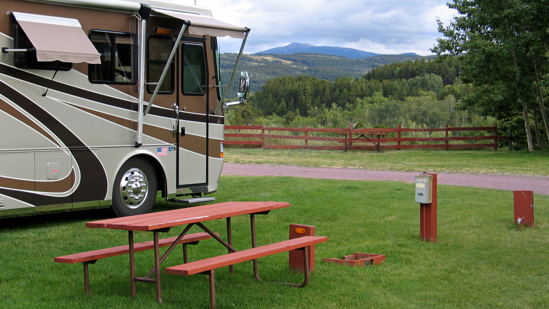 RV parked at a campsite with a picnic table, power hookup, and view of a wooden fence and mountains in the background.
