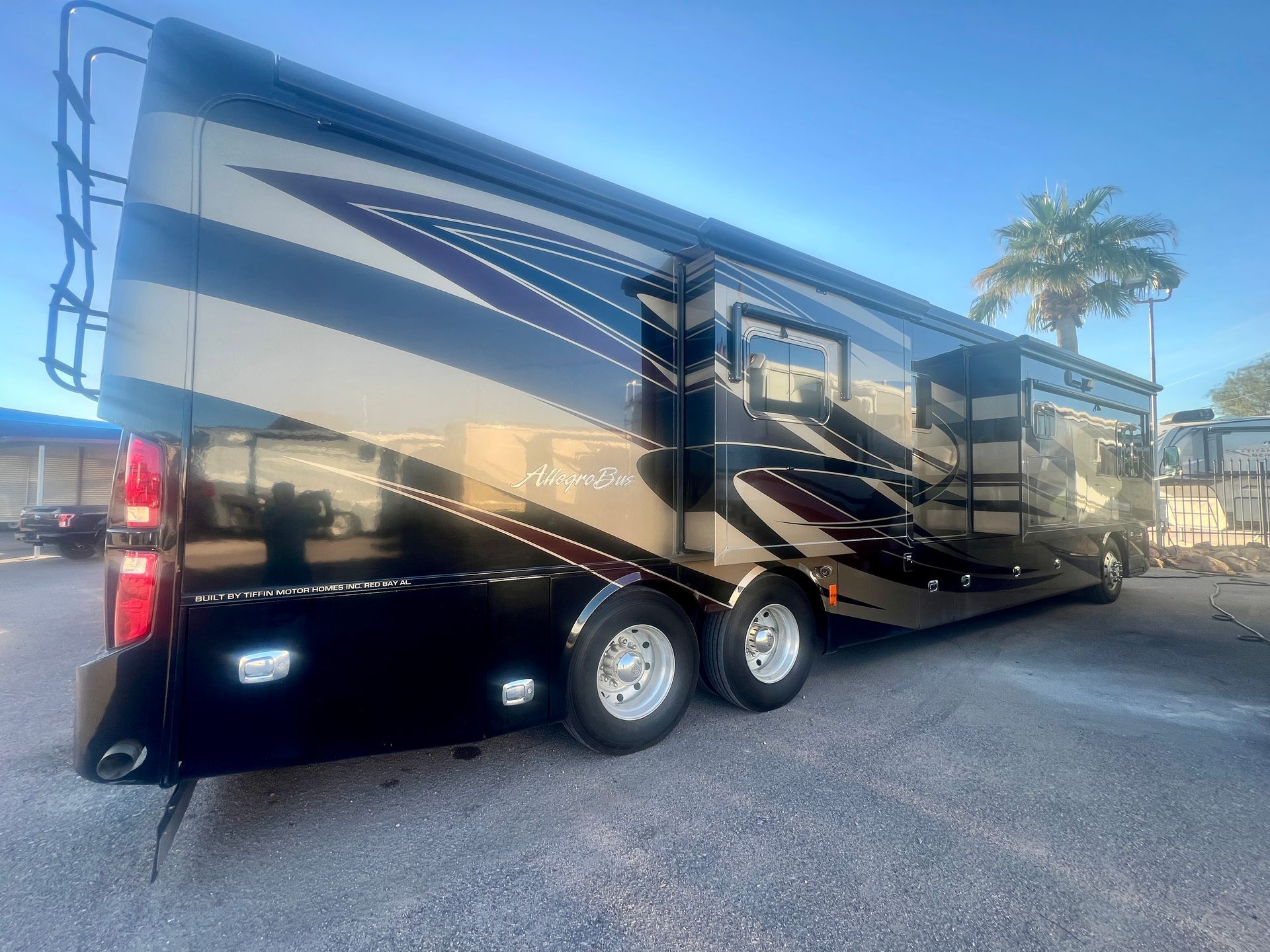 Black and tan RV parked outdoors on a sunny day; ladder on the back.
