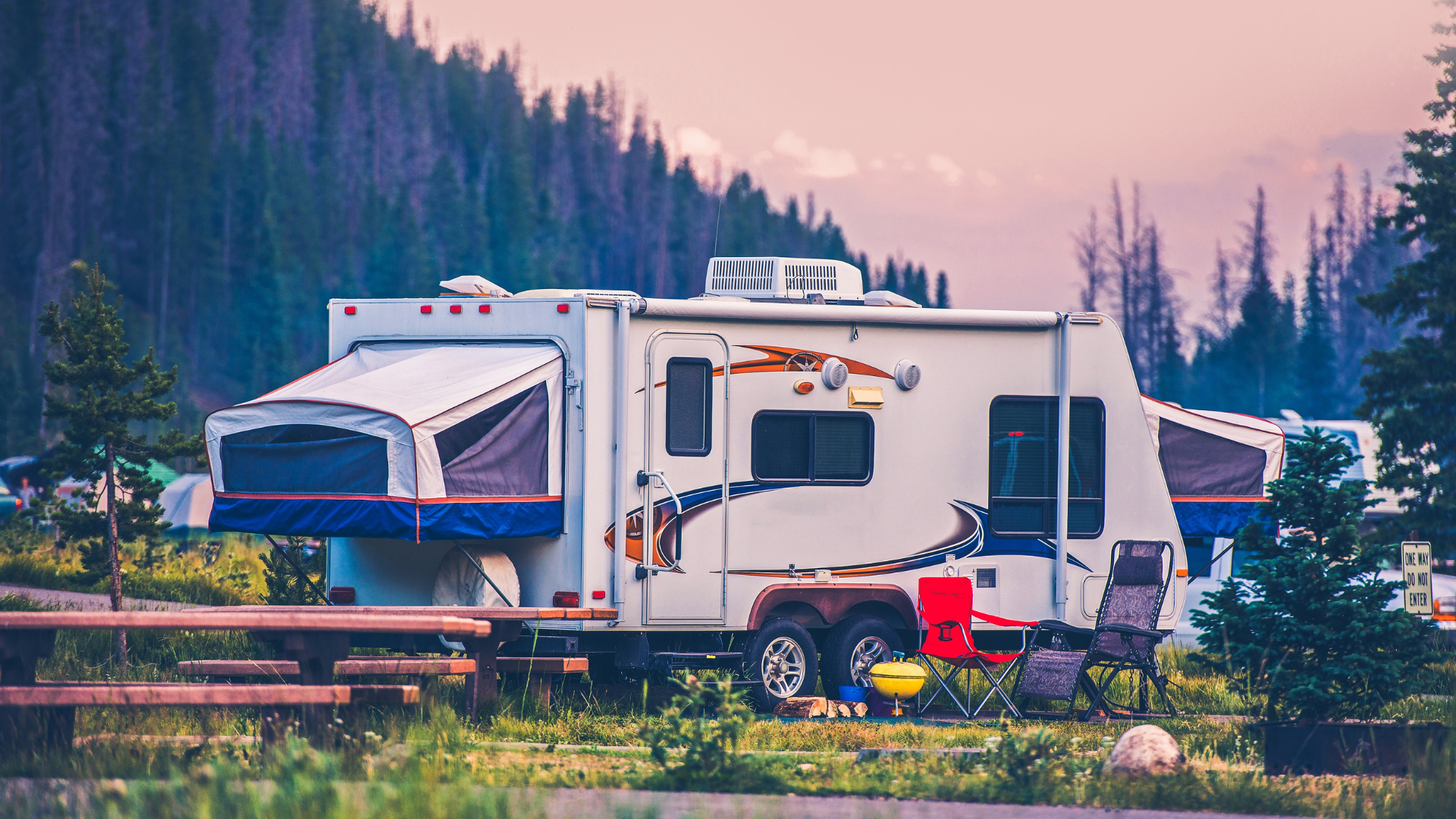 RV with pop-out bed in a campsite. White and blue camper, with picnic table and forested mountain backdrop.