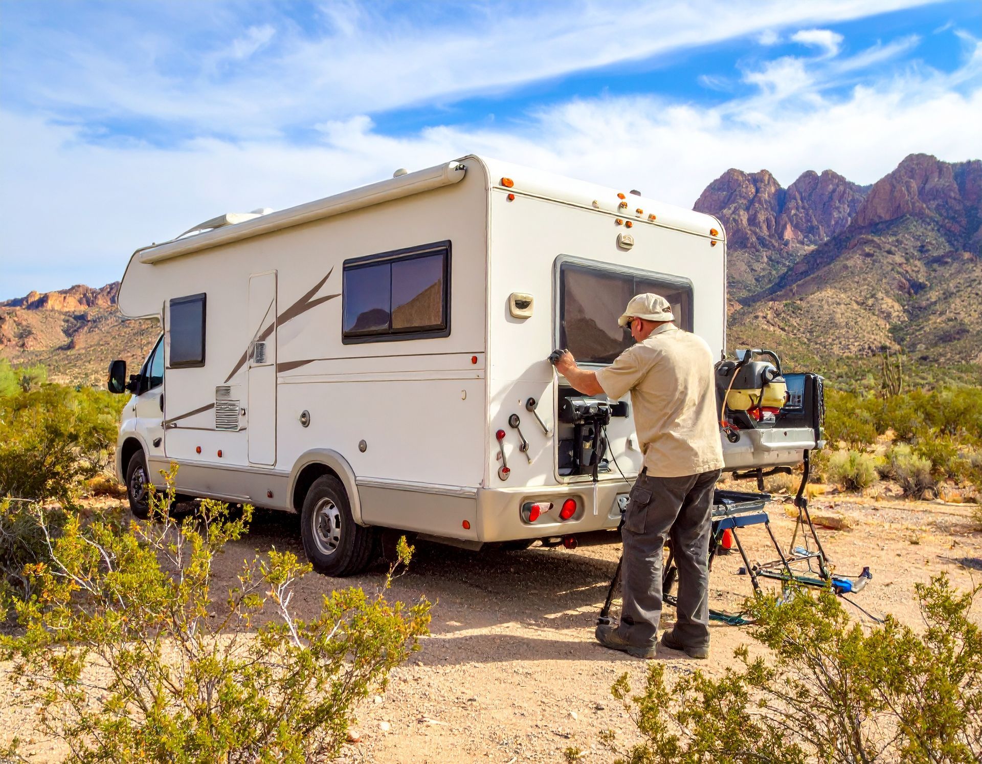 A man adjusts equipment near the back of a white RV parked in a desert landscape with mountains in the background.