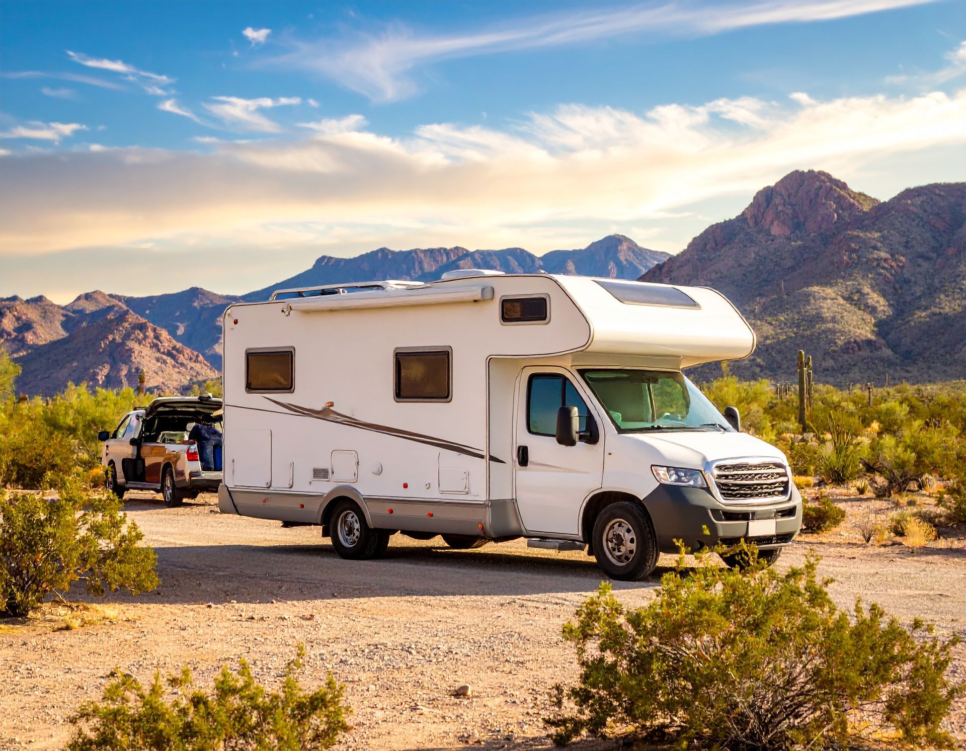 White RV and towing vehicle parked on a dirt road in a desert landscape, with mountains in the background under a blue sky.