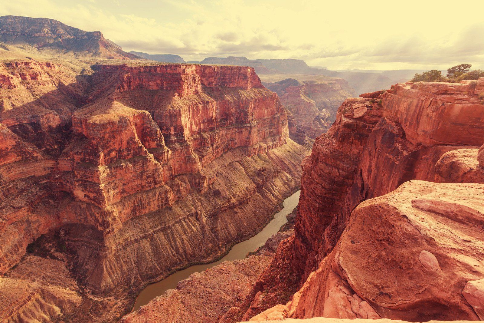 Grand Canyon vista. Red and brown rock formations surround a winding river, seen under a yellow-toned sky.