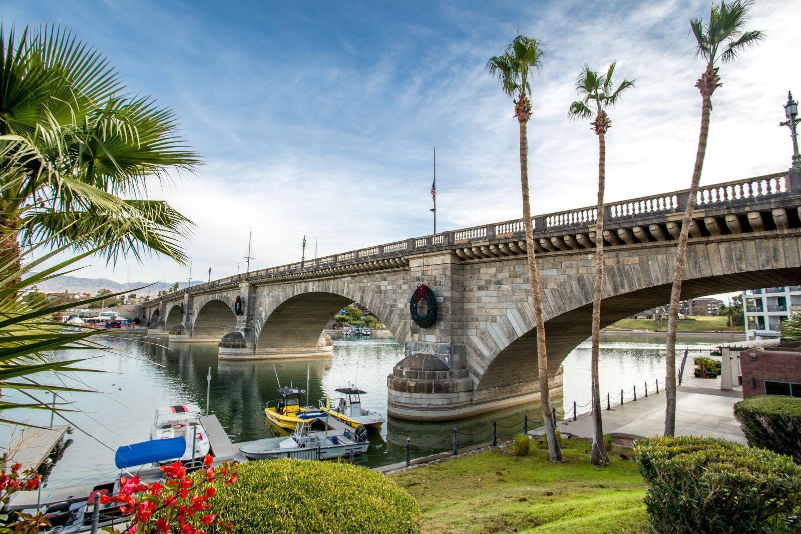 London Bridge spanning a waterway, palm trees in the foreground, boats docked below, and clear blue sky.