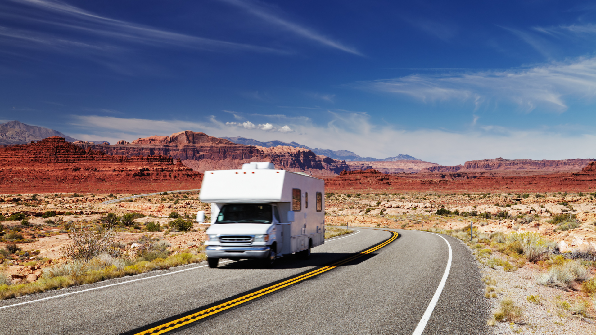 RV driving on a desert highway with red rock formations and a blue sky.