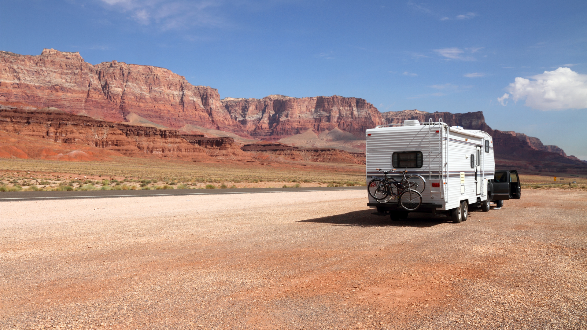 RV parked on a dirt road with a red rock mountain range in the background under a blue sky.