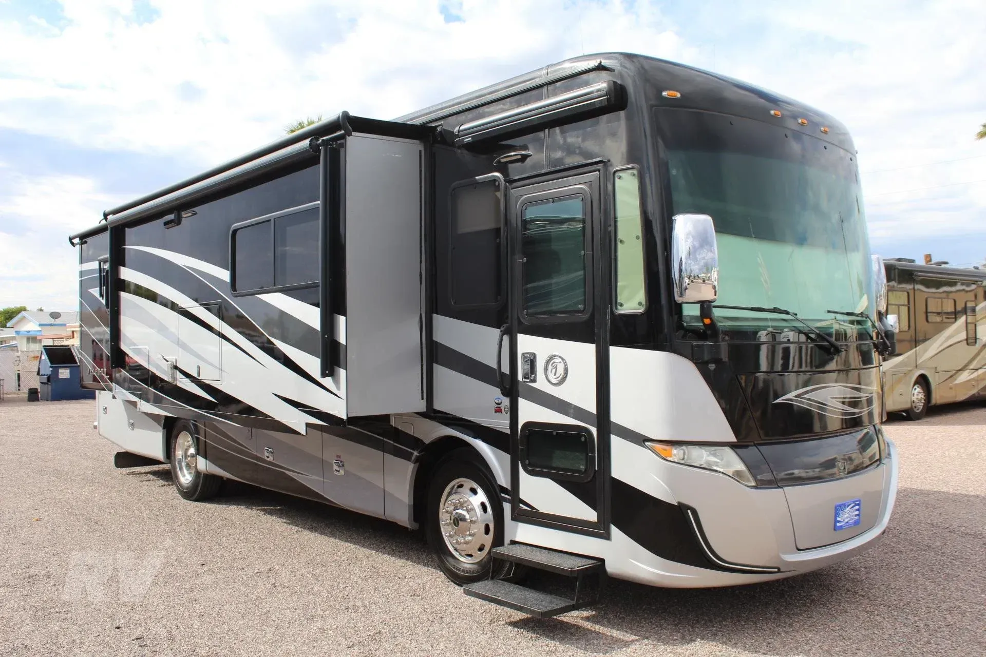 Large black and silver recreational vehicle with slide-out extended, parked on gravel.