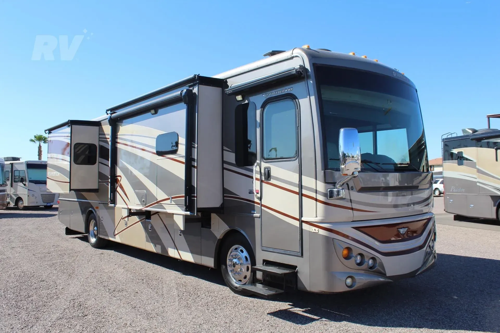 Tan and brown RV with slide-outs extended, parked on gravel in bright sunlight.
