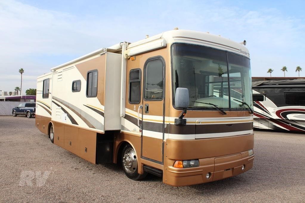 Brown and white RV parked on gravel, with a slide-out extended and palms in the background.
