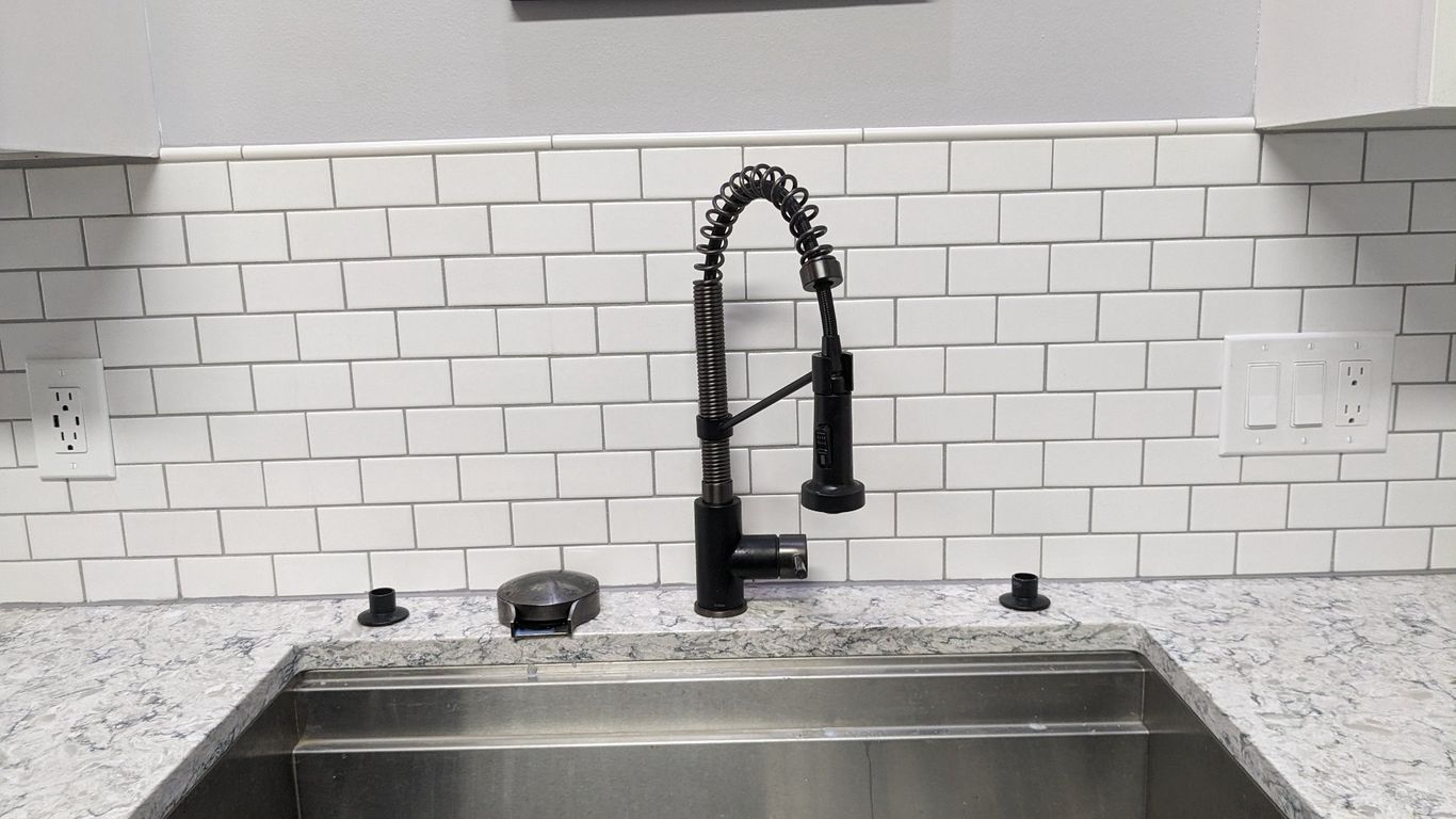 A black kitchen faucet with a pull-down sprayer sits above a stainless steel sink against a white subway tile backsplash.