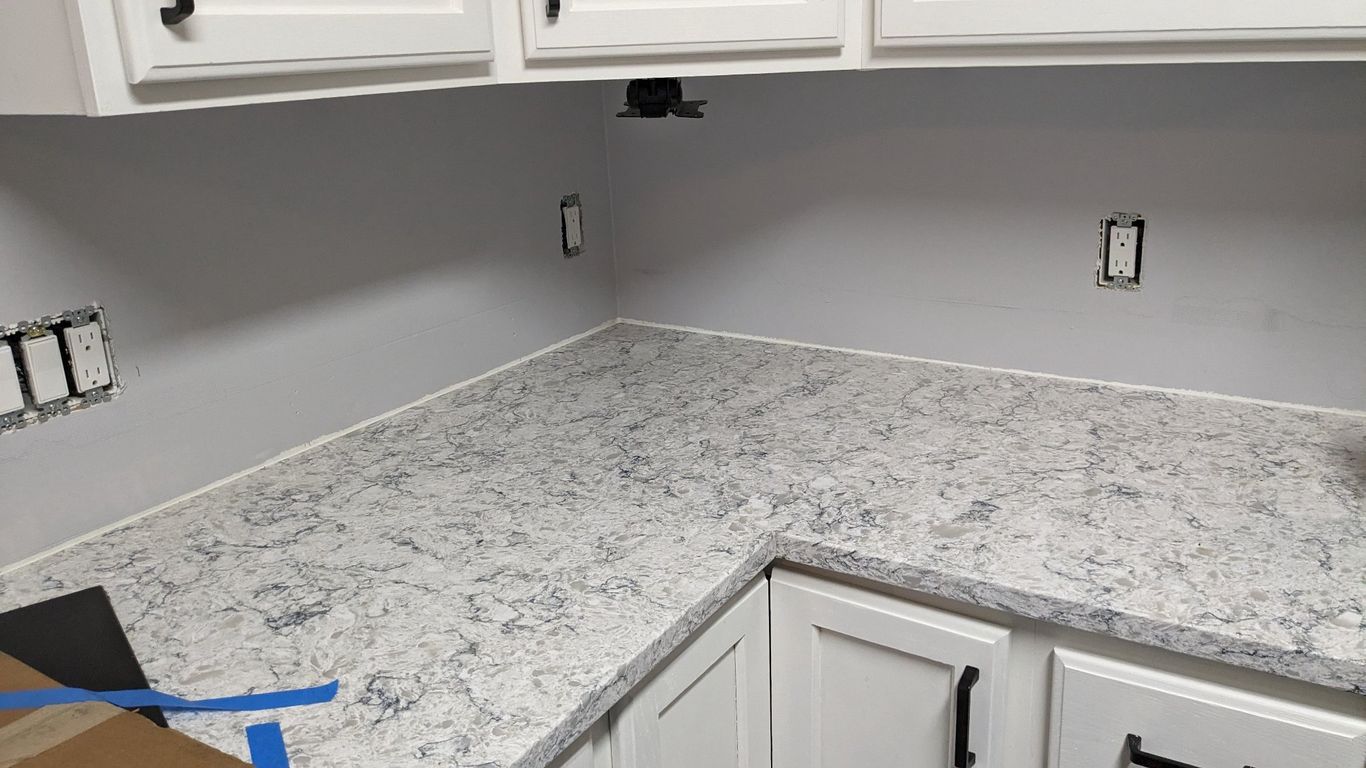 White cabinets and speckled countertop in a kitchen corner with electrical outlets and gray walls.