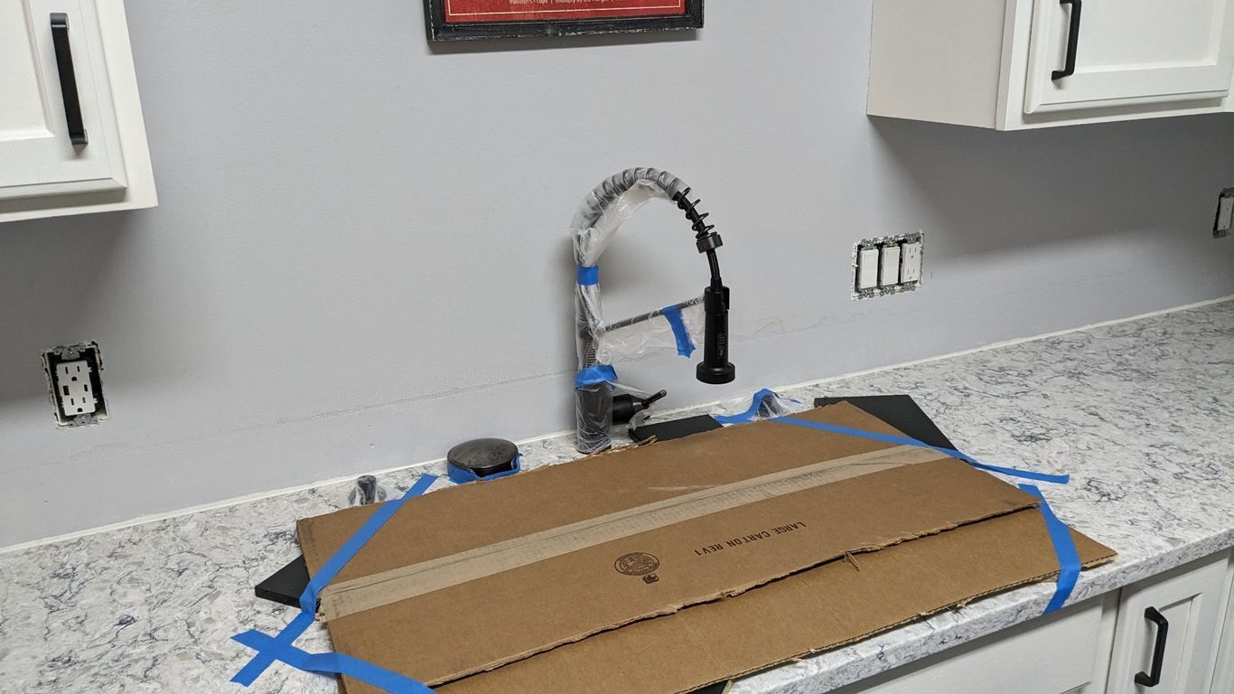 Kitchen with sink, faucet, and countertop protected by cardboard, blue tape. Gray walls, white cabinets.