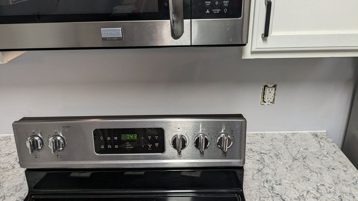 Stainless steel oven and microwave in a kitchen, with a grey backsplash and power outlet.