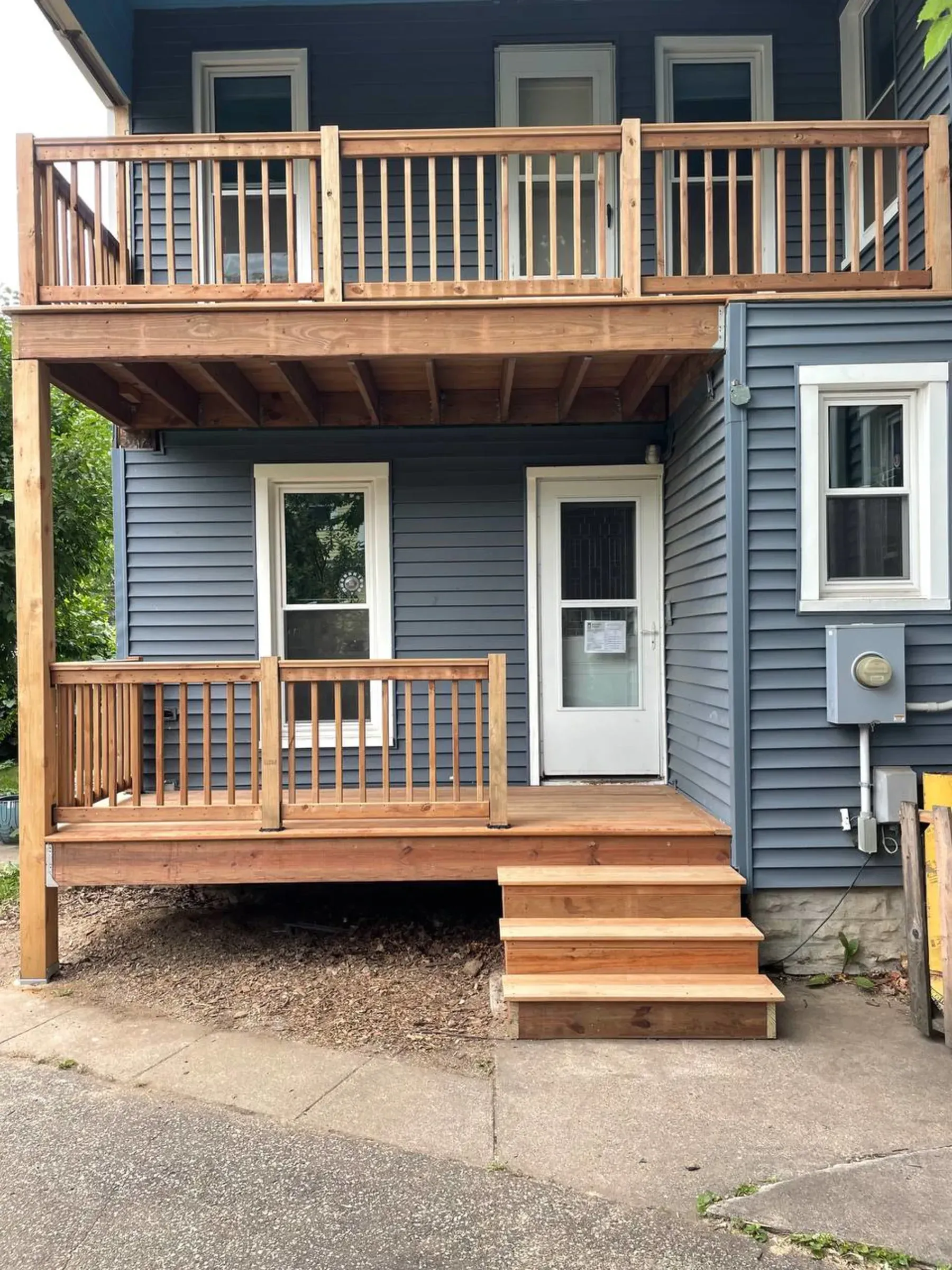 Two-story building exterior with a wooden deck and stairs. Blue siding, white door and windows, brown wood railing.