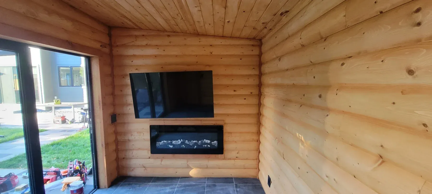 Interior of a wooden cabin with a TV above a fireplace. Large glass door on the left, natural lighting.