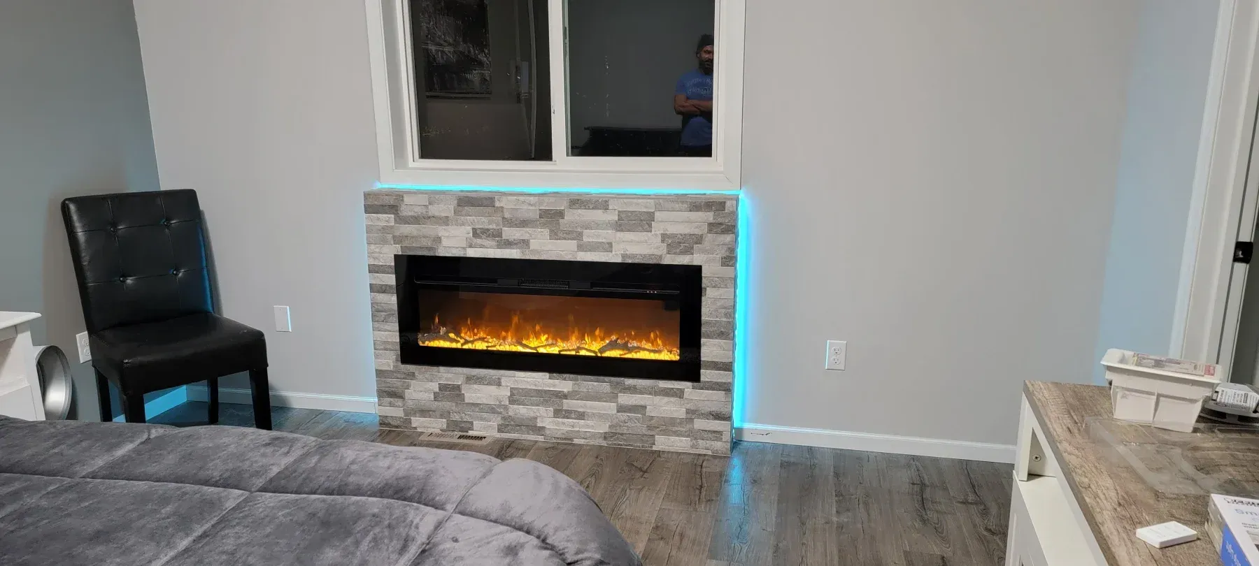 Bedroom with an electric fireplace with a stone facade under a window, next to a leather chair.