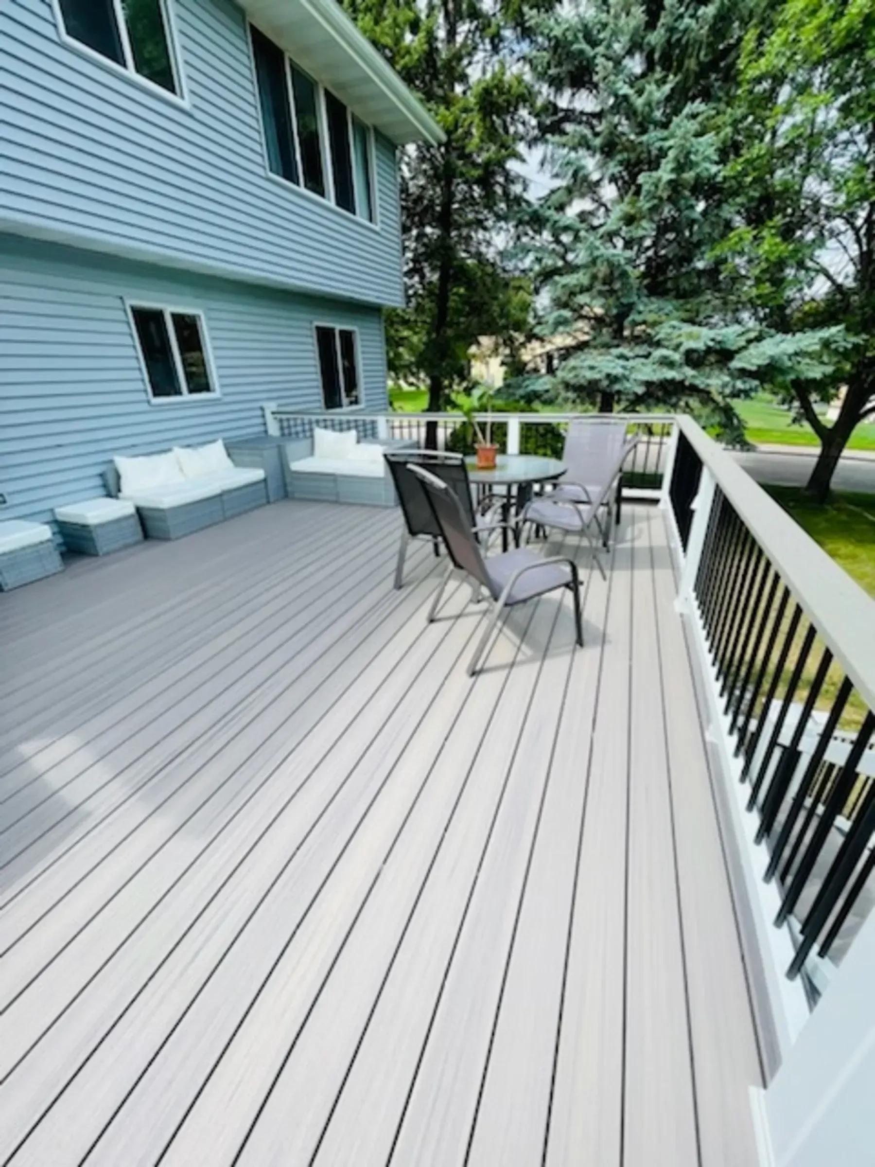 A spacious deck with gray flooring and black railing, furnished with seating and a table, alongside a blue house.