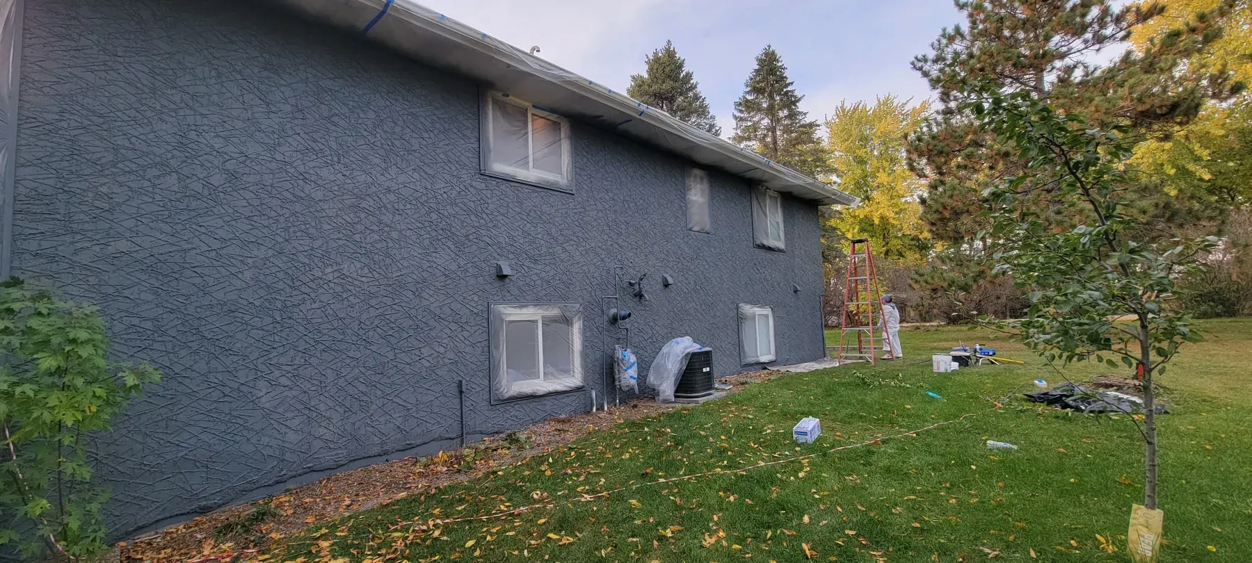 Blue building with white-framed windows, surrounded by green grass and trees. Overcast sky.