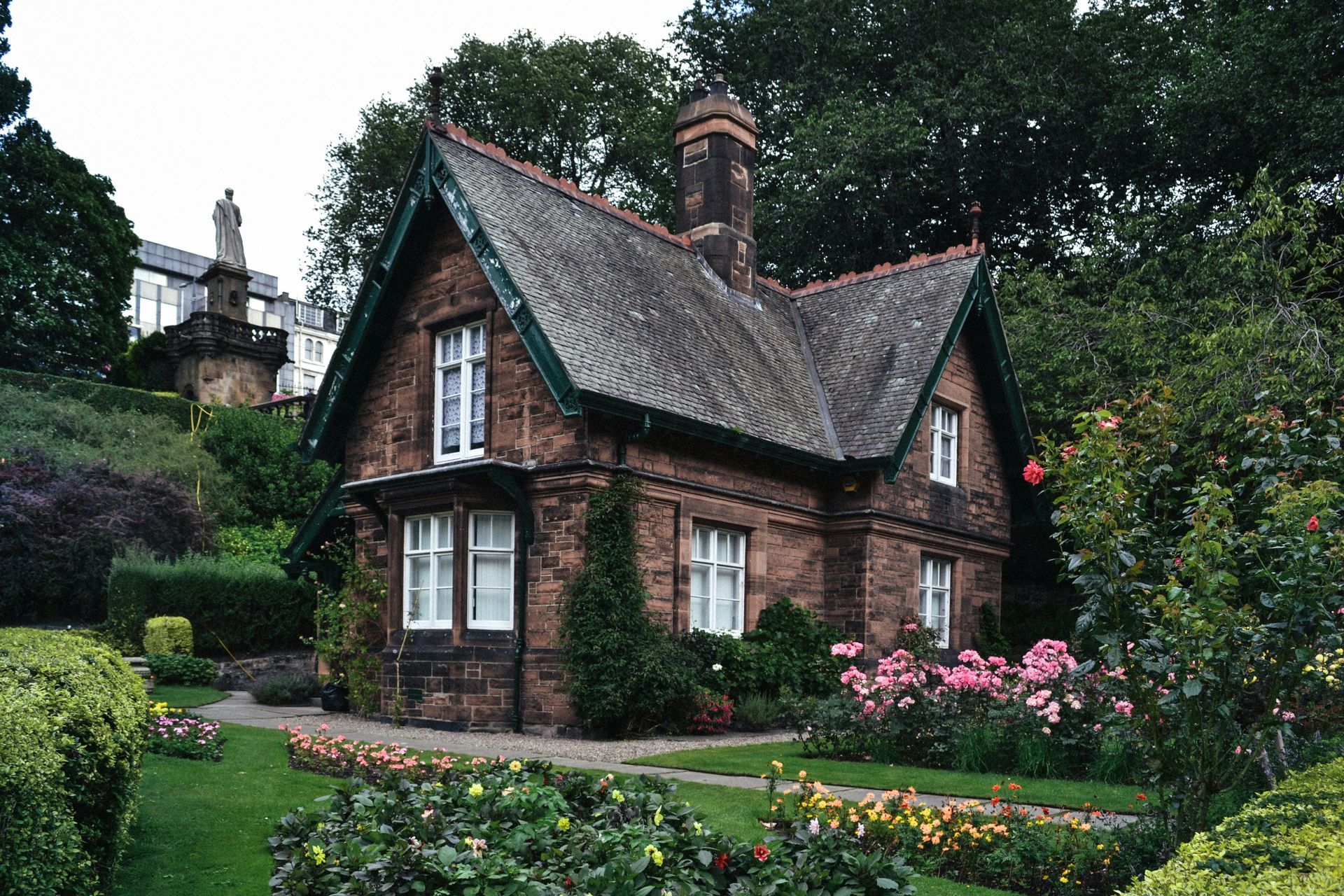 A small brick house with a green roof is surrounded by flowers and bushes.