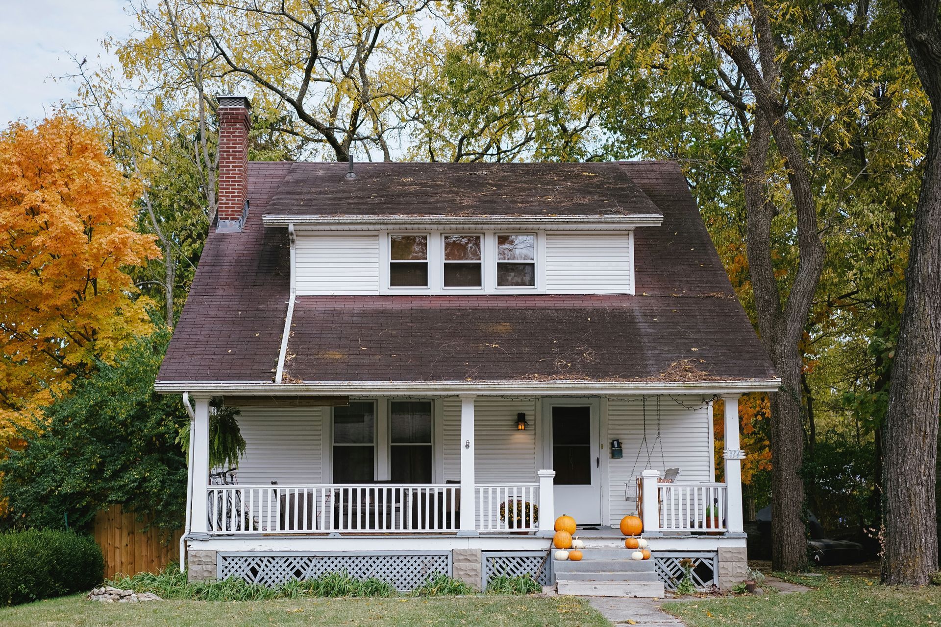 A white house with a porch and pumpkins on it is surrounded by trees.