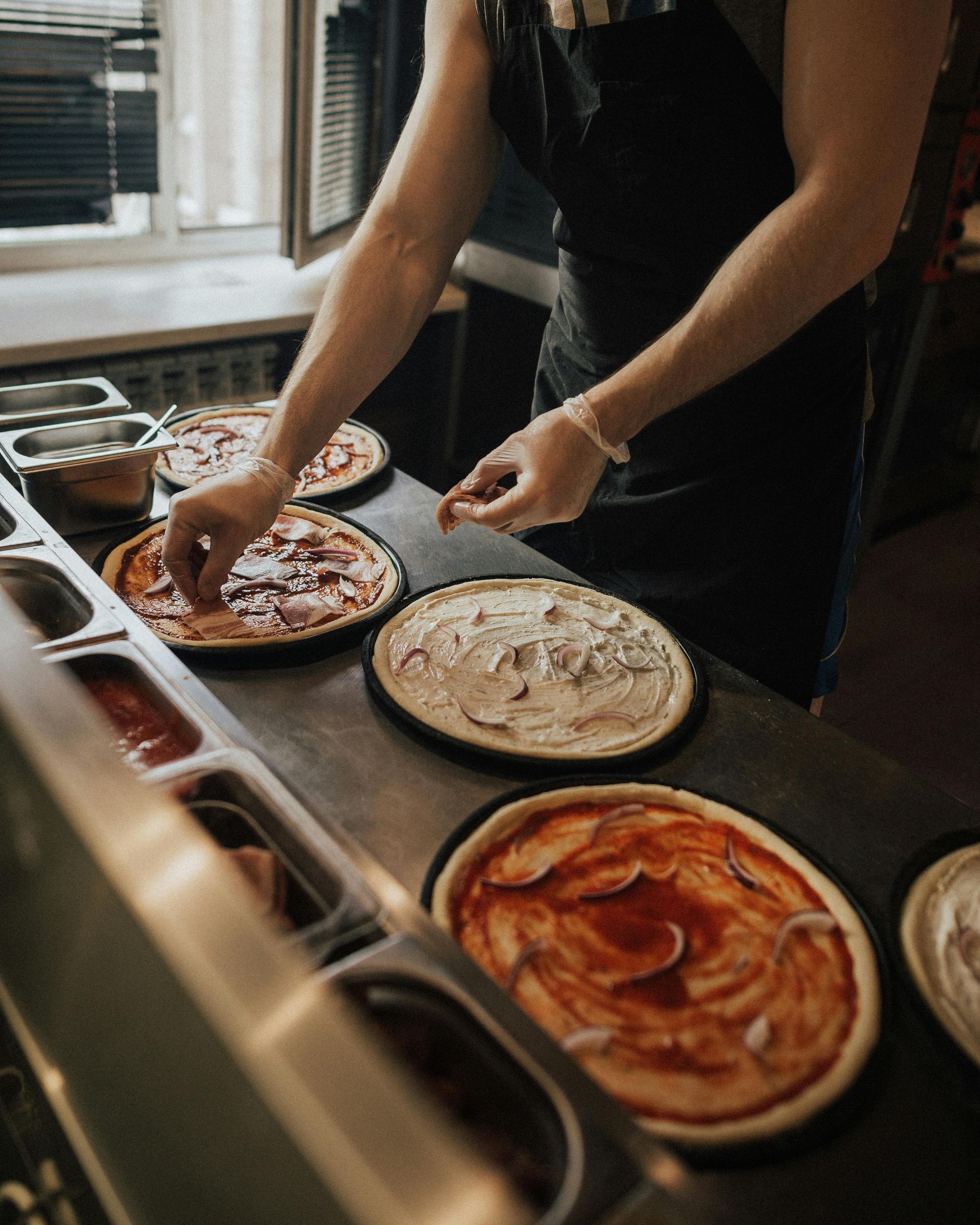 Persona che prepara le pizze nella cucina di un ristorante, preparando i condimenti per diverse pizze.
