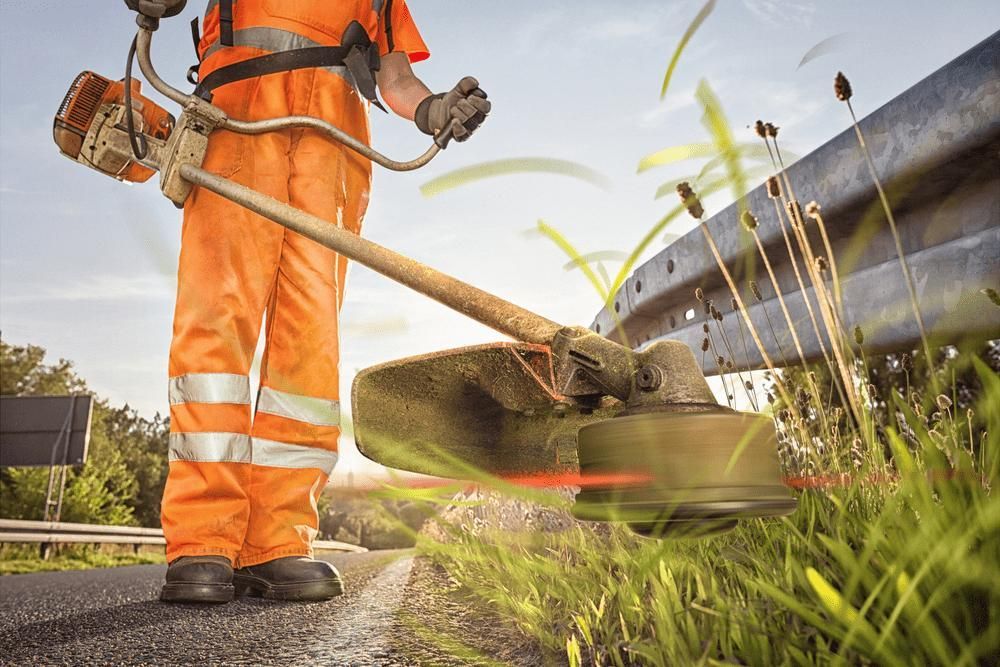 A Man Is Using A Lawn Mower To Cut Grass On The Side Of The Road — Leslie Farm & Garden Machinery In South Grafton, NSW