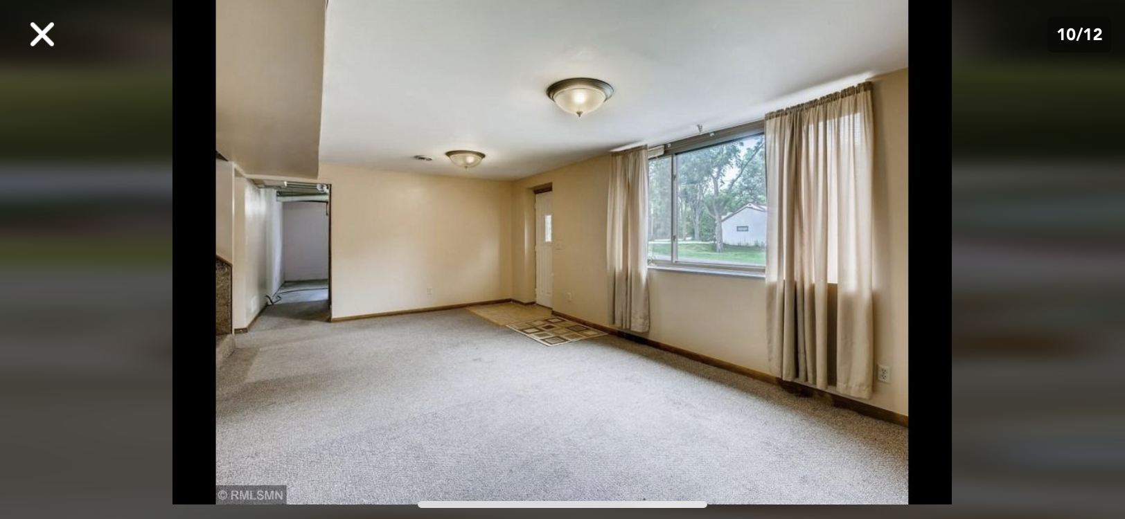 An empty room with light beige walls, gray carpeting, two ceiling lights, and a window with floor-length curtains.